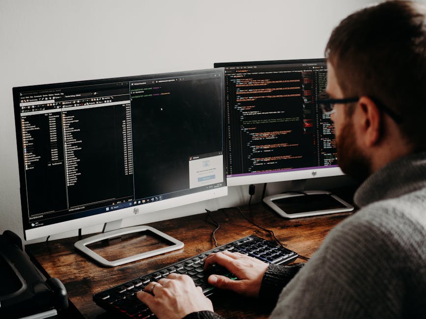 The image shows a person working at a desk with two large computer monitors displaying lines of code, suggesting a programming or software development activity. The individual, wearing glasses and a g