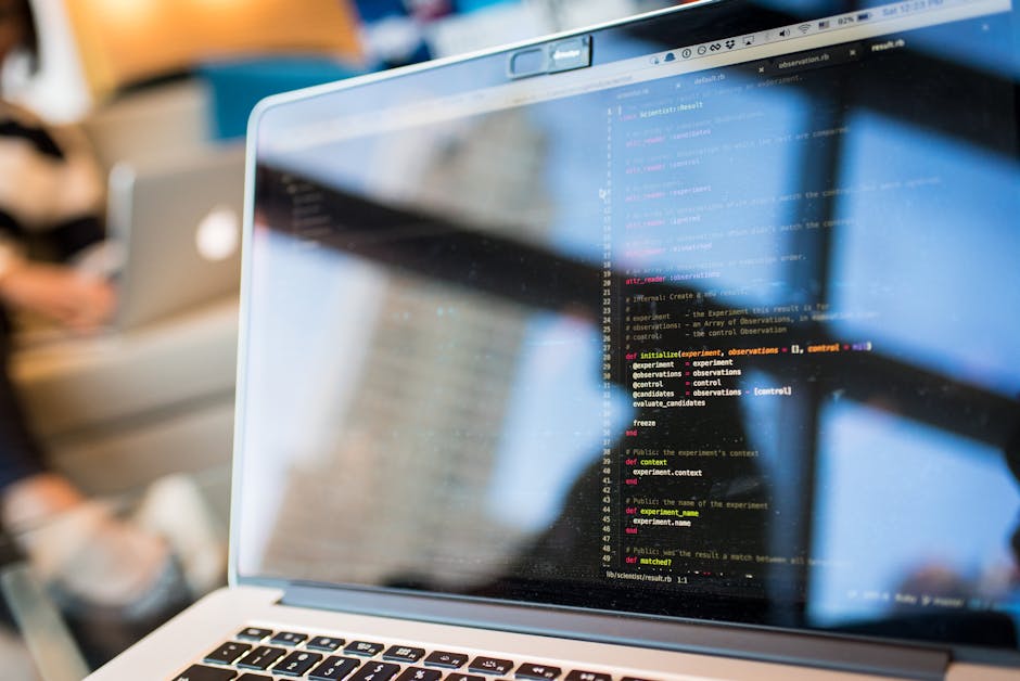 The image shows a person working at a desk with two large computer monitors displaying lines of code, suggesting a programming or software development activity. The individual, wearing glasses and a g